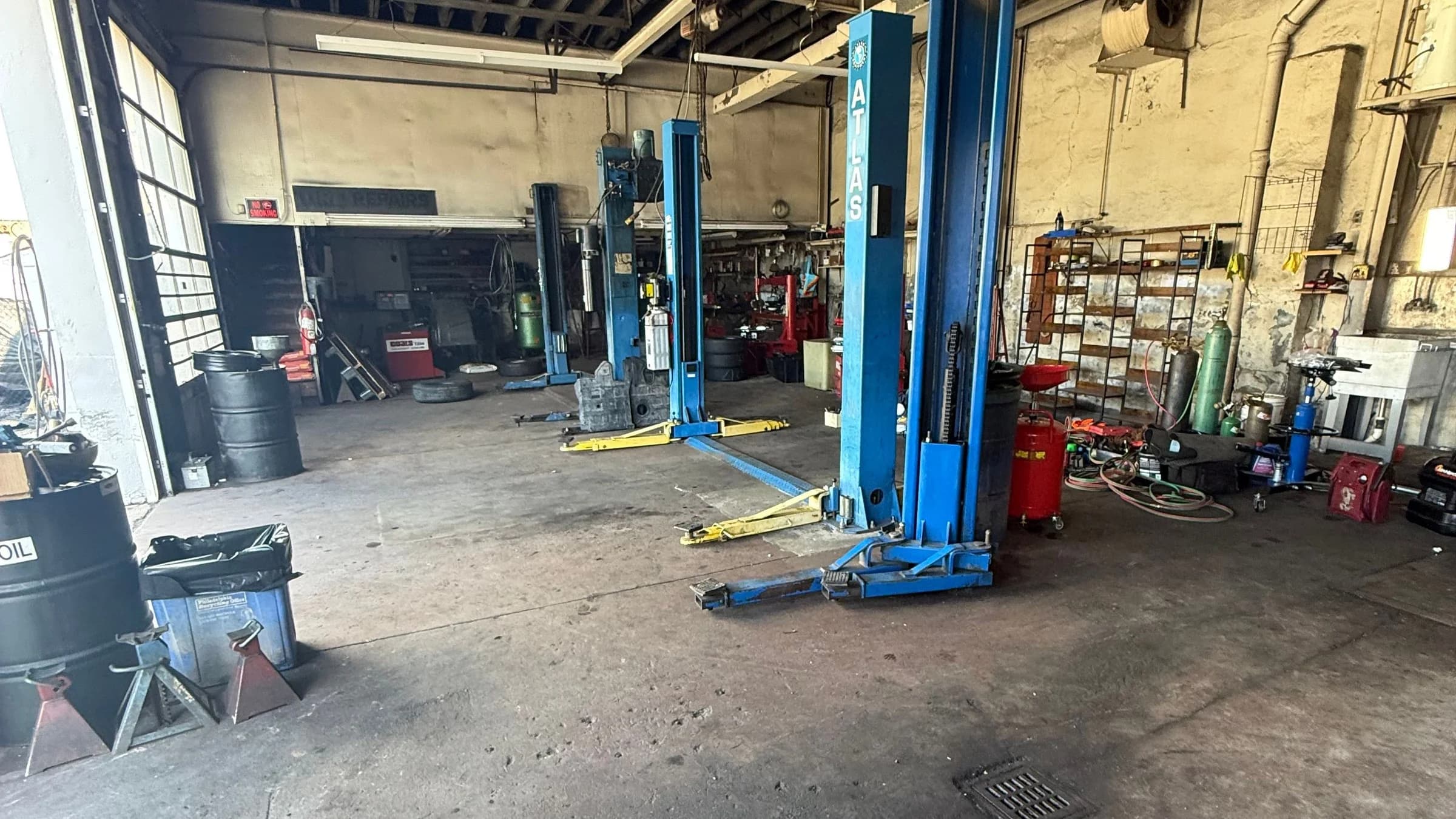 Inside AlongSide Garages — two-post lifts on a clean concrete floor, tools and oil barrels along the walls, daylight from the open bay door.