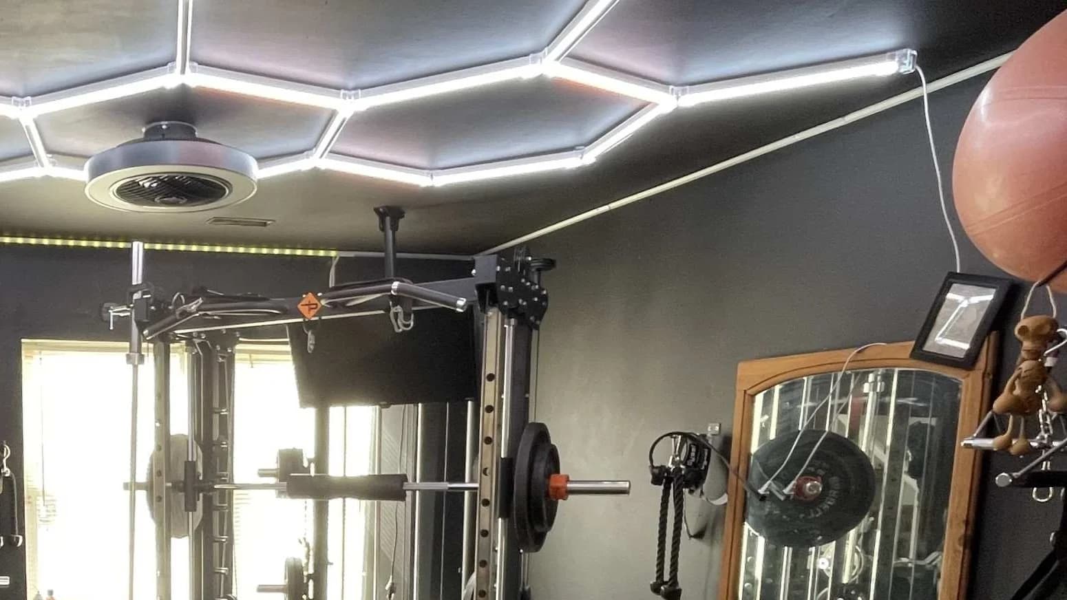 A home gym with a power rack, pull-up bar, cable pulley attachment, loaded barbell, and an adjustable bench, lit by a hexagonal LED ceiling panel.